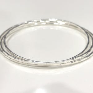 Op de afbeelding: Drie zilveren armbanden met een gehamerde textuur, liggend op een wit oppervlak. De armbanden zijn cirkelvormig en hebben een licht onregelmatige vorm, die het licht reflecteert. De sieraden zijn eenvoudig en elegant.