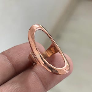Pode incluir: Um anel em tom de cobre com uma estrutura grande em formato oval alongado. A superf&iacute;cie met&aacute;lica apresenta um acabamento martelado e texturizado com um brilho quente. O design consiste numa banda fina e cont&iacute;nua em forma de la&ccedil;o aberto.