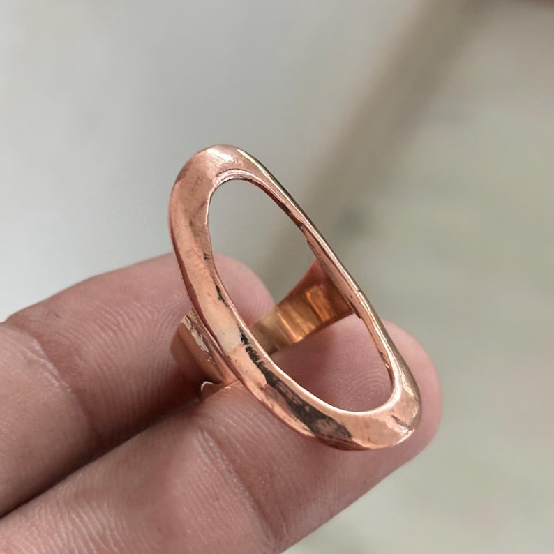Pode incluir: Um anel em tom de cobre com uma estrutura grande em formato oval alongado. A superf&iacute;cie met&aacute;lica apresenta um acabamento martelado e texturizado com um brilho quente. O design consiste numa banda fina e cont&iacute;nua em forma de la&ccedil;o aberto.