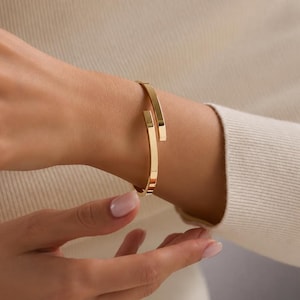 Può includere: Un bracciale rigido color oro lucido caratterizzato da un design aperto con due estremità piatte e parallele che si sovrappongono leggermente. Il bracciale presenta una superficie liscia e riflettente con una struttura rigida attorno al polso.