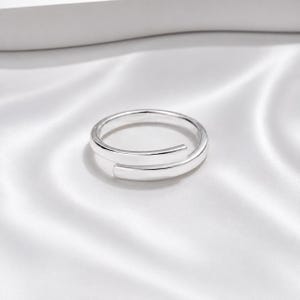 May include: A polished silver-tone bypass ring featuring a smooth, continuous band that overlaps at the front. The ring has a reflective metallic finish and a simple, open-ended design that allows for slight adjustment in fit around the finger.