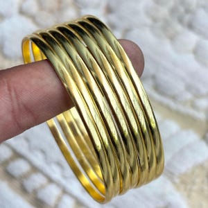 Può includere: Un set di sette braccialetti color oro. Ogni braccialetto ha un esterno liscio e arrotondato con una sottile texture a coste. I braccialetti sono impilati, creando un effetto a strati. I gioielli sono tenuti da un dito.