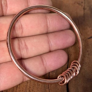 May include: A copper-colored bangle bracelet with a simple, circular design. The bracelet features a series of small, connected rings at one point. The jewelry is held in a hand, showcasing the warm, metallic hue of the bracelet.