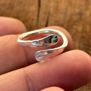 Könnte beinhalten: Ein silberfarbener Wickelring mit gehämmertem Muster. Der Ring hat ein offenes Design, das sich um den Finger wickelt. Das Metall reflektiert das Licht und erzeugt ein glänzendes Aussehen. Der Ring wird auf einem Finger gezeigt.