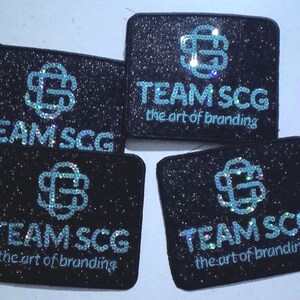 Puede incluir: Cuatro parches rectangulares negros con acabado brillante. Cada parche presenta un logotipo entrelazado de color azul claro sobre el texto "TEAM SCG" y el texto más pequeño "the art of branding" en azul claro, todo con un estilo centelleante.