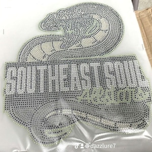 Pode incluir: Um aplique de strass com um design de cobra enrolada em tons de preto e prata. O texto SOUTHEAST SOUL ALLSTARS está integrado no gráfico com uma fonte em bloco em negrito. Todo o design é composto por pequenas pedras refletoras brilhantes.