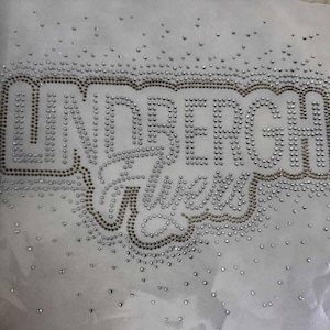 May include: White fabric featuring the text "UNDERBERG" and "Alles" in silver and gold rhinestones. The rhinestones are scattered across the fabric, creating a sparkling effect.