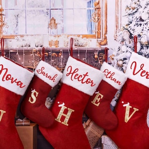 Puede incluir: Cinco calcetines navideños rojos con ribete de piel sintética blanca, cada uno personalizado con un nombre en escritura roja. Letras y detalles dorados adornan los calcetines, que cuelgan frente a un fondo festivo.