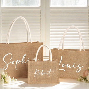 Puede incluir: Tres bolsas de mano de yute con asas blancas y nombres personalizados en fuente cursiva. La bolsa m&aacute;s grande dice "Sophia", la mediana "Louis" y la m&aacute;s peque&ntilde;a "Robert". Las bolsas est&aacute;n colocadas frente a una ventana con flores.