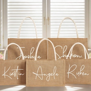 May include: A collection of burlap tote bags with white handles and personalized names in elegant script. The bags vary in size and are arranged in front of a window with soft lighting, suggesting a wedding or event theme.