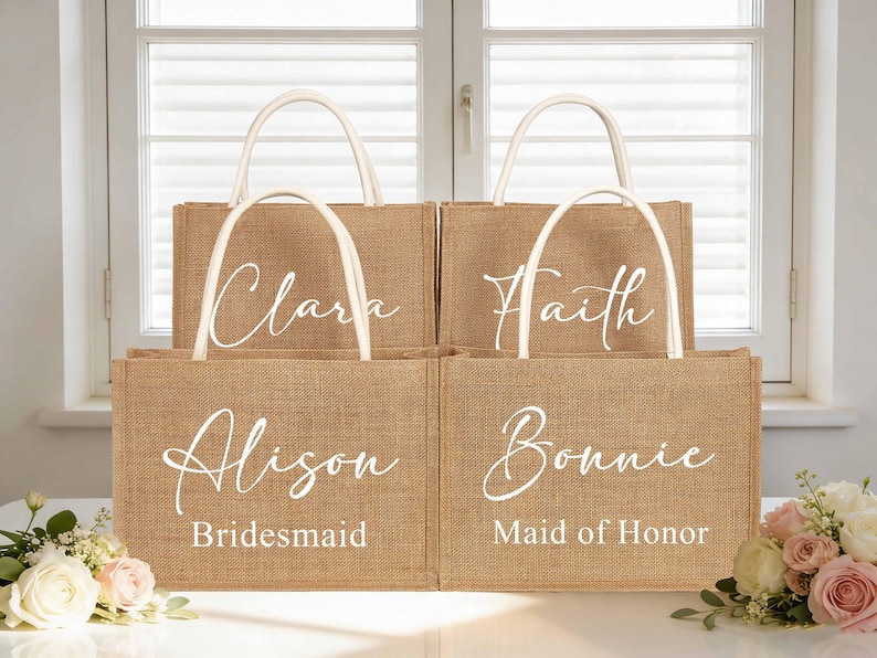 May include: Four burlap tote bags with white handles and personalized names in elegant script. The bags are labeled "Clara", "Faith", "Alison - Bridesmaid", and "Bonnie - Maid of Honor". The bags are arranged in front of a window.