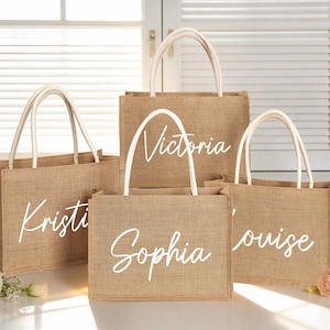 May include: Four jute tote bags with white handles, each bearing a name in white script: "Kristi", "Victoria", "Sophia", and "Louise". The bags are displayed in front of a window with white blinds, with floral arrangements in the foreground.