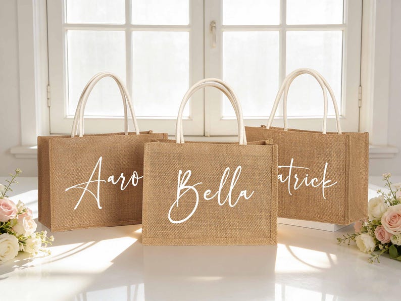 Puede incluir: Tres bolsas de yute con asas blancas, cada una con un nombre en escritura blanca: Aaron, Bella y Patrick. Las bolsas est&aacute;n colocadas sobre una superficie blanca, con una ventana detr&aacute;s y decoraciones florales a ambos lados.