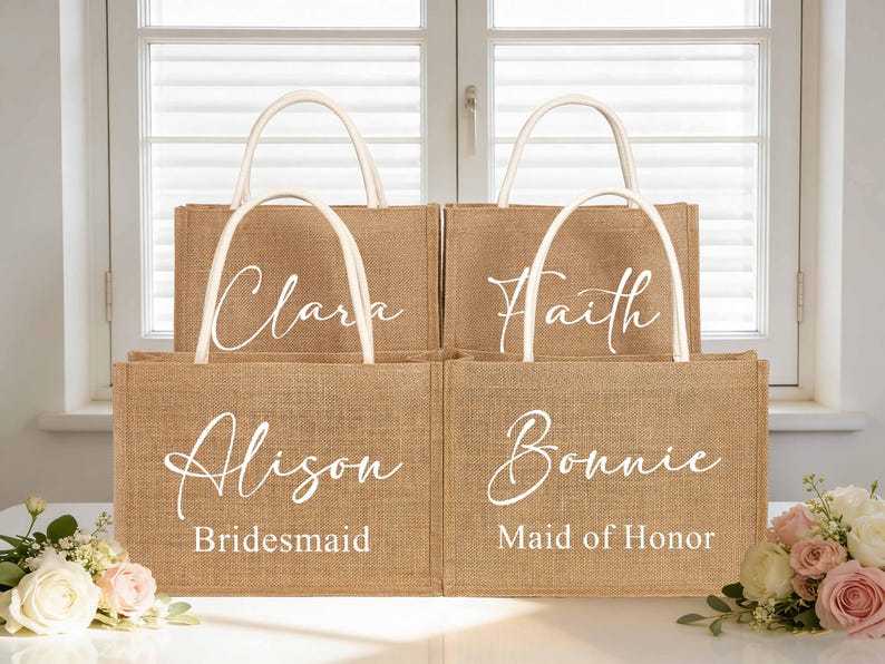 Puede incluir: Cuatro bolsas de yute con asas blancas, cada una con un nombre en escritura elegante. Las bolsas est&aacute;n etiquetadas como "Clara", "Faith", "Alison - Bridesmaid" y "Bonnie - Maid of Honor". Est&aacute;n dispuestas frente a una ventana.