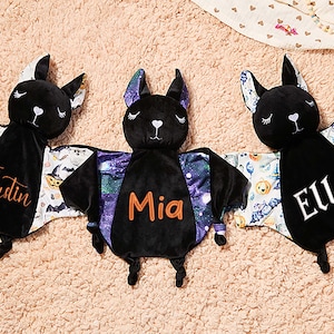 Puede incluir: Tres peluches negros con forma de murciélago con nombres personalizados en naranja o blanco. Los murciélagos tienen alas estampadas con diseños temáticos de Halloween. Los nombres "Justin", "Mia" y "Ella" se muestran.