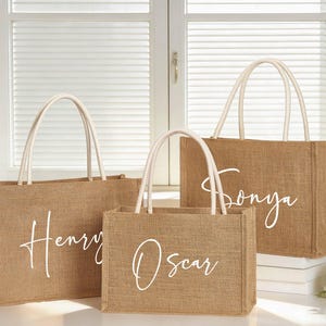 Puede incluir: Tres bolsas de mano de yute con asas blancas y nombres personalizados: "Henry", "Oscar" y "Sonya" en fuente cursiva. Las bolsas son rectangulares y de color marr&oacute;n natural.