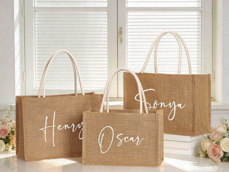 Puede incluir: Tres bolsas de mano de yute con asas blancas y nombres personalizados: "Henry", "Oscar" y "Sonya" en fuente cursiva. Las bolsas son rectangulares y de color marr&oacute;n natural.
