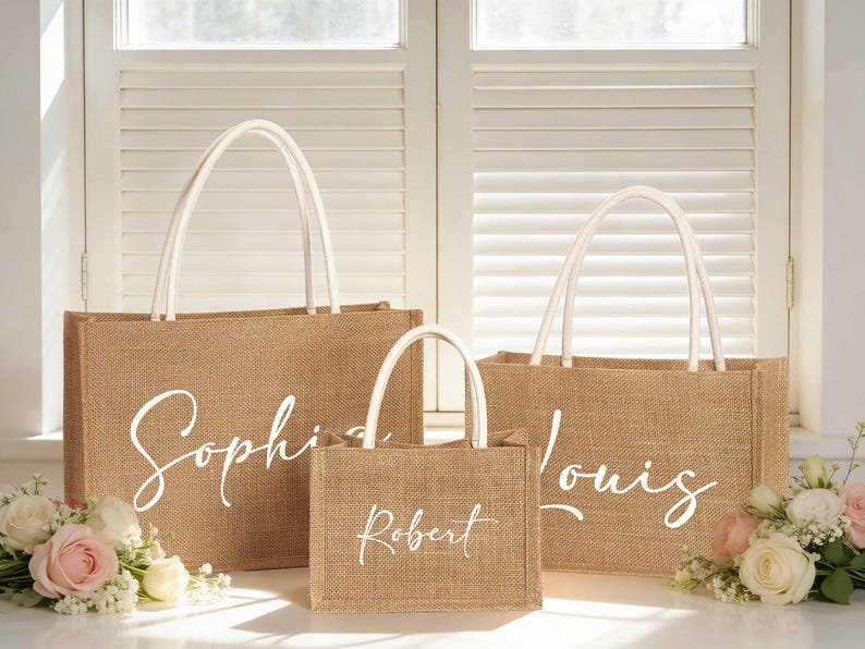 May include: Three burlap tote bags with white handles and custom names in a cursive font. The largest bag says "Sophia", the medium bag says "Louis", and the smallest bag says "Robert". The bags are sitting in front of a window with flowers.
