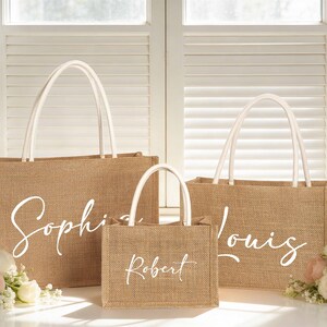 May include: Three burlap tote bags with white handles and custom names in a cursive font. The largest bag says "Sophia", the medium bag says "Louis", and the smallest bag says "Robert". The bags are sitting in front of a window with flowers.