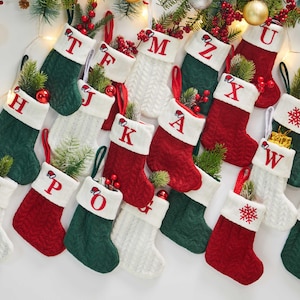 Op de afbeelding: Een verzameling kerstsokken in rood, groen en wit, elk met een letter geborduurd op de manchet. Sommige sokken hebben een sneeuwvlokontwerp. De sokken zijn gevuld met groen en bessen en hangen met kerstversieringen.