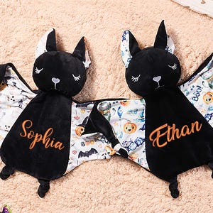 May include: Two black bat-shaped baby security blankets. Each has a black head with embroidered eyes and a heart-shaped nose. The wings are patterned with Halloween-themed images. One blanket is embroidered with the name "Sophia", the other with "Ethan".
