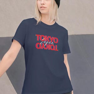 Puede incluir: Camiseta azul marino con el texto "TOKYO Japan TOKYO" en rojo y blanco. El diseño está centrado en la parte delantera. La camiseta es de manga corta.