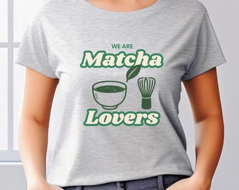 以下が含まれることがあります： 「WE ARE Matcha Lovers」の文字が緑と白でプリントされたライトグレーのTシャツ。抹茶のボウル、泡立て器、葉っぱのデザイン。クルーネックのTシャツです。