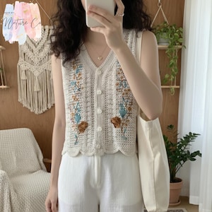 Pode incluir: Um colete de crochê sem mangas creme com decote em V e fecho de botão. O colete apresenta bordados florais em azul, castanho e amarelo. A bainha inferior tem uma borda ondulada. Uma sacola branca é visível.