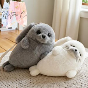 May include: Two plush seal toys, one gray and one white, are displayed on a textured surface. The gray seal has a strap and a zipper. The white seal is lying down with its eyes closed. The text "MADE BY: Nature Co" is in the upper left corner.