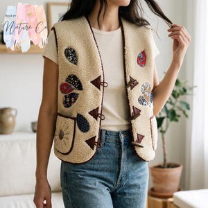 Puede incluir: Chaleco de forro polar color crema con parches decorativos con motivos florales y geométricos. El chaleco presenta ribetes marrones oscuros, cierres de palanca y un bolsillo con un diseño floral. El texto "MADE BY. Nature Co" está en la esquina superior izquierda.
