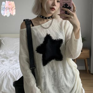 May include: Off-white, long-sleeved sweater with a large, black star design. The sweater has an off-the-shoulder neckline. A black leather shoulder bag is visible. The image also shows a black choker necklace with silver chain and a phone.