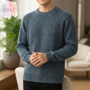 Puede incluir: Un suéter azul texturizado de cuello redondo con un pequeño emblema dorado "Y" en el pecho. El suéter tiene un patrón sutil y una pequeña etiqueta en la parte inferior izquierda. También se ve el texto "MADE BY NATURE CO".