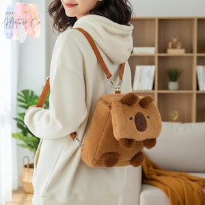 May include: A brown plush backpack shaped like a capybara, with brown ears, feet, and a face. The backpack has brown straps and silver metal clasps. The backpack is being worn by a person wearing a cream-colored hoodie.