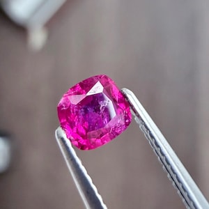 以下が含まれることがあります： 銀色のピンセットで保持された、鮮やかなクッションカットのルビーの宝石。 ルビーは、光を反射する内部ファセットを備えた、深いピンクがかった赤色を示しています。 宝石の形状は、丸みを帯びた角を持つ正方形です。