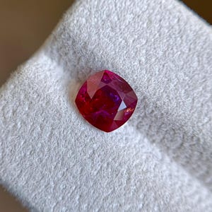 May include: A cushion-cut gemstone with a deep, vibrant red color and multiple facets that reflect light. The stone displays a translucent quality with internal inclusions visible throughout its structure, showcasing a rich, saturated crimson hue.