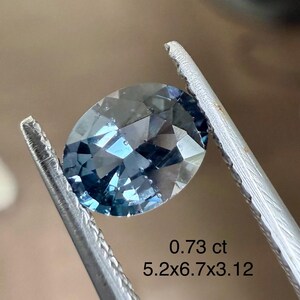 May include: An oval-cut, blue gemstone held by tweezers. The gem has a faceted surface and a slight blue hue. Text on the image reads "0.73 ct" and "5.2x6.7x3.12".