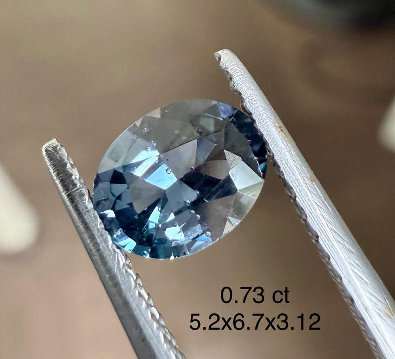 May include: An oval-cut, blue gemstone held by tweezers. The gem has a faceted surface and a slight blue hue. Text on the image reads "0.73 ct" and "5.2x6.7x3.12".