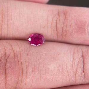 May include: A round, faceted, deep red gemstone. The gem is held in a hand, highlighting its brilliant cut and rich colour. The stone's facets reflect light, creating a sparkling effect. This gemstone could be a ruby or similar precious stone.