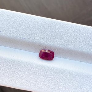 Op de afbeelding: Een gefacetteerde, rechthoekige robijn edelsteen met een dieprode kleur. De edelsteen ligt op een witte, getextureerde ondergrond, die de gepolijste facetten en rijke kleur benadrukt. De robijn heeft een kussensnede.