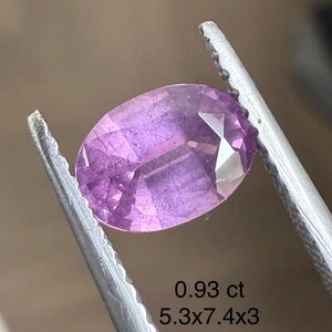 May include: A faceted, oval-cut, purplish-pink gemstone held by tweezers. The gem weighs 0.93 ct and measures 5.3 x 7.4 x 3 mm. The gem's facets reflect light, showcasing its clarity and color. The background is a neutral tone.