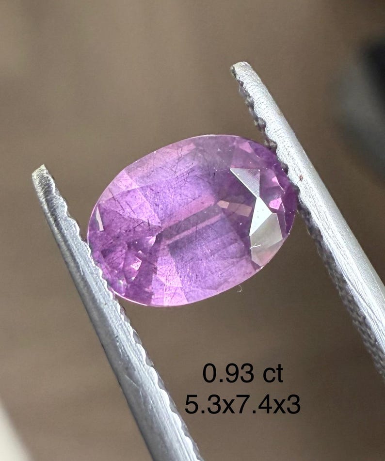 May include: A faceted, oval-cut, purplish-pink gemstone held by tweezers. The gem weighs 0.93 ct and measures 5.3 x 7.4 x 3 mm. The gem's facets reflect light, showcasing its clarity and color. The background is a neutral tone.