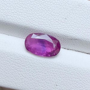 以下が含まれることがあります： 鮮やかなマゼンタと紫の色合いを持つ、ファセットカットされた楕円形の宝石。石の表面は研磨されており、光を反射する複数の幾何学的なファセットが施されています。深く飽和した色合いと滑らかな輪郭が特徴です。