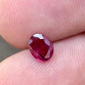 May include: A faceted, oval-shaped ruby gemstone. The ruby is a deep, rich red color with visible internal inclusions. The gemstone is held between two fingers, showcasing its size and brilliance. The cut of the ruby allows light to reflect, creating a sparkling effect.