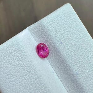 May include: A vibrant, oval-cut, pink gemstone. The gem is set against a white, textured surface. The gemstone's facets reflect light, creating a sparkling effect. This gemstone is suitable for jewelry making.