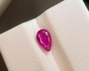 Puede incluir: Una gema de rubí facetada en forma de pera. La gema es de un color rojo rosado intenso y vibrante, y descansa sobre una superficie blanca texturizada. El corte de la piedra es visible, mostrando sus facetas.