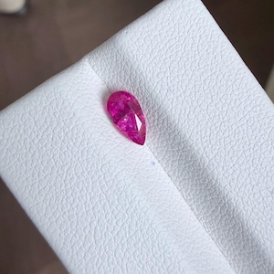 Puede incluir: Una gema rosa brillante en forma de pera y facetada. La gema está colocada sobre una superficie blanca y texturizada. El corte de la piedra permite que la luz se refleje, creando un efecto brillante. La gema es probablemente para joyería.