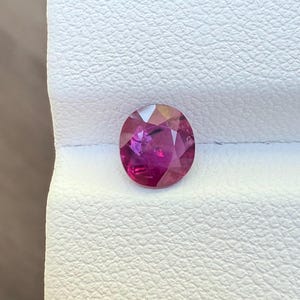 May include: A round, faceted gemstone with a deep pinkish-red hue. The gem is set against a white textured surface, showcasing its brilliance and clarity. This gemstone could be used in jewelry.