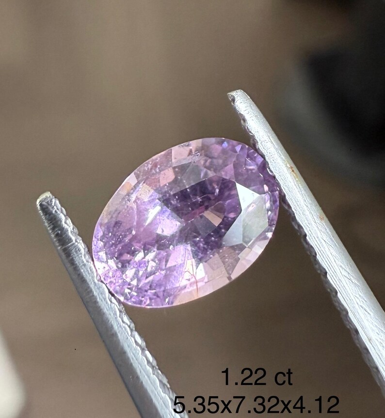 May include: A faceted, oval-shaped, light purple gemstone is held by a pair of silver tweezers. The gem is 5.35 x 7.32 x 4.12 mm and weighs 1.22 ct. The gem has a brilliant cut and is a light lavender color.