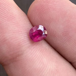May include: A faceted, oval-shaped ruby gemstone. The ruby is a deep, vibrant red color with multiple facets that reflect light. The gemstone is held between two fingers, showcasing its size and brilliance.