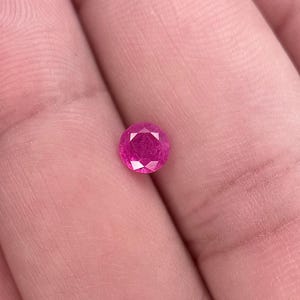 Puede incluir: Una gema de rubí redonda y facetada. La gema es de un vibrante tono rojo rosado, con múltiples facetas que crean un efecto brillante. La gema se sostiene en una mano, mostrando su tamaño y color.